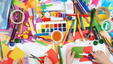 An assortment of craft supplies, paints, markers and hands with scissors cutting paper.