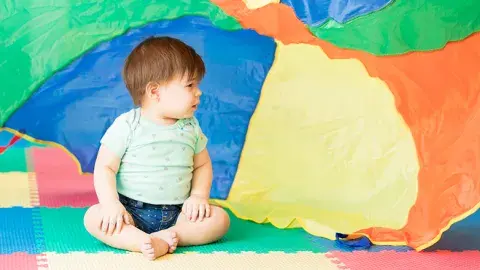 A baby sits on a colorful mat, with a colorful cloth waving behind them.
