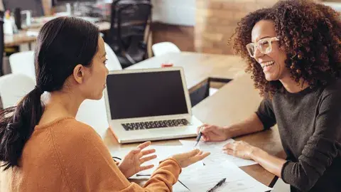 Two women sitting at a table with a laptop and papers.