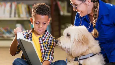 A young boy sitting in a library holds open a book for a white, shaggy dog while an encouraging woman looks on. 
