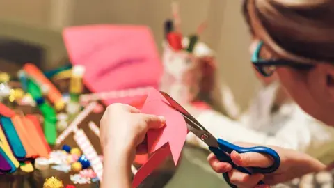A woman cuts pink paper, art supplies are on a table in front of her.
