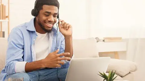 a young African-American man smiling sitting on a couch, looking down at a laptop that's on a small table, with headphones on
