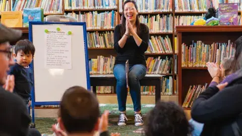 A woman sits in front of a group of toddlers and adults clapping her hands. A white board is next to her facing the crowd. 
