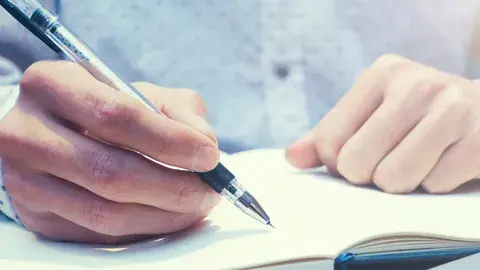 Close-up of two hands resting on an open journal, with the right hand holding a pen to write.