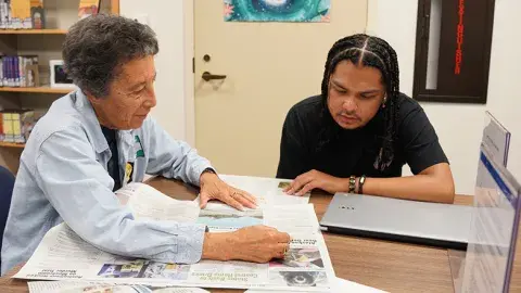 A person helping another person read the newspaper.