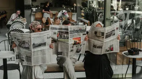 Three ladies sitting in chairs in a row holding up newspapers in front of their faces