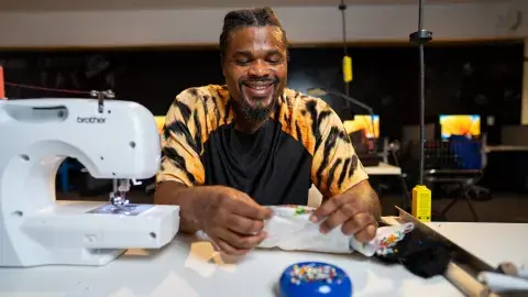A man in tie-died shirt smiles while he works on a textile project at a white table with a sewing machine. 