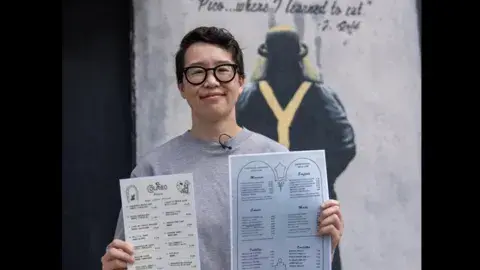 person holds up two different menus in front of muralled exterior wall