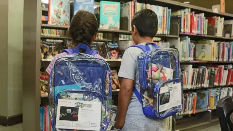 Two children wear clear backpacks filled with play kits.