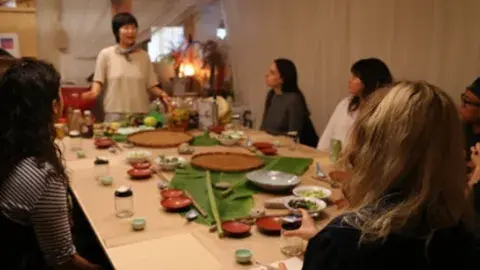 Picture of Jessica Wang giving a food preparation demo to people seated around a table.