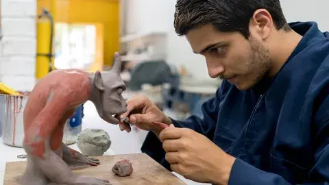 A man sculpts a fanciful creature from clay using pottery tools.
