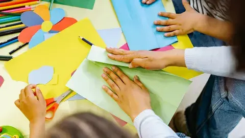 An assortment of craft supplies, paints, markers and hands with scissors cutting paper.