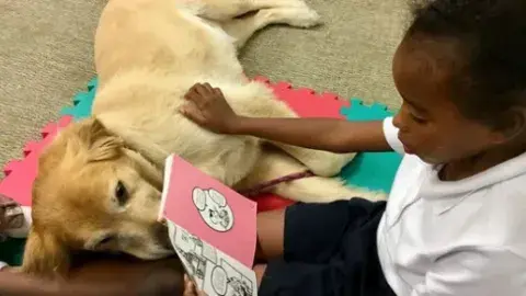 A young girl reading to a dog who is lying down with its head in her lap.