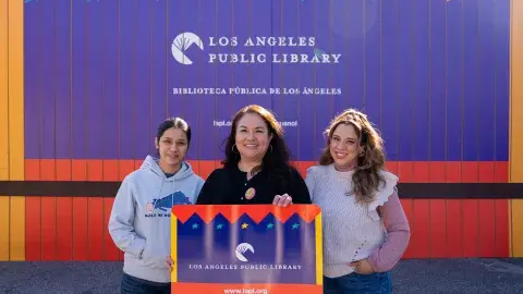 2 librarians and a clerk holding a giant library card stand in front of the temporary building