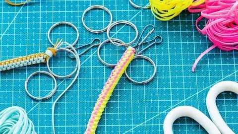 ​The image shows a boondoggle keychain surrounded by all the craft supplies used to make it such as multi-colored gimp string, scissors, and scattered key rings. 