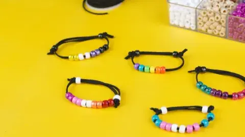 Colorful beaded bracelets on black cords are arranged on a vibrant yellow surface. A box of assorted beads is partially visible in the background.