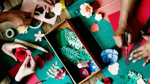 Three pairs of hands use crafting tools to make paper flowers.
