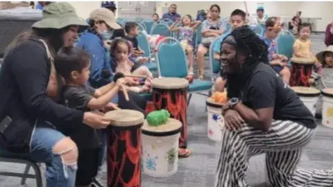 Woman kneeling in front of a mother and child playing a drum