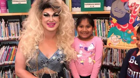 A smiling drag queen in a blonde wig poses in the library with two smiling children