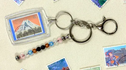 A keychain made of a postage stamp and beads is surrounded by a scattering of postage stamps.