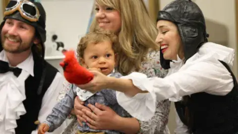 Image is of two LA Opera performers in costume holding a bird prop in front of a young mother with her baby toddler.