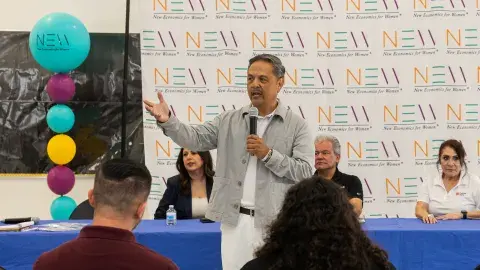 A man holding a microphone gestures in front of an audience with seated panelists behind him.