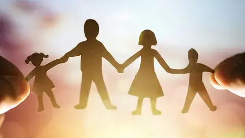 Silhouette of a paperdoll family being held by two hands against a bright background.
