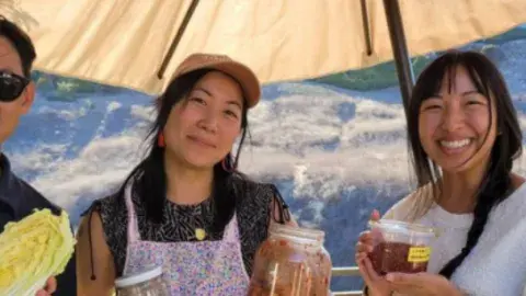3 people are pictured holding jars of pickles under an umbrella