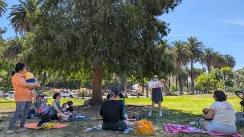 librarian reads a picturebook to a group of adult caregivers and toddlers on a grass lawn under a shady tree