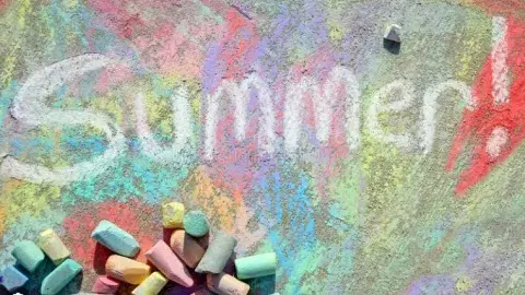Colorful sidewalk chalk art with the word “Summer!” written on pavement beside scattered chalk pieces.