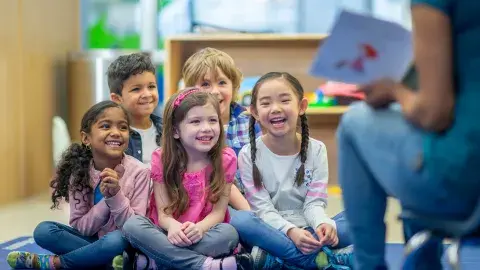 A group of children are smiling and laughing while listening to a Storyteller read a book. 