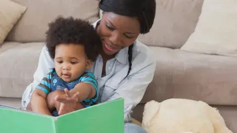 A mother reads from a book to the baby sitting in her lap.