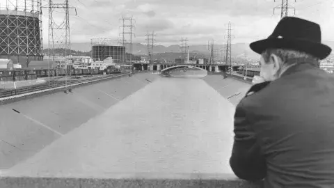 Person in overcoat and wide-brimmed hat facing away from the camera, looking down at the Los Angeles River with industrial tanks in the background.
