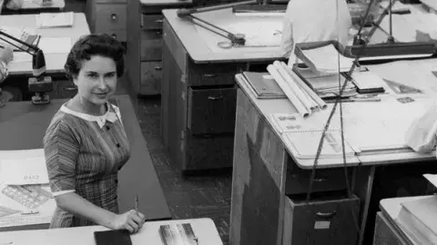 Newspaper employee standing while other employees sit and work at desks.
