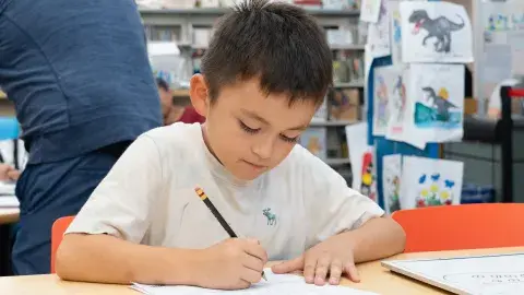 A young boy sits at a table in a library and writes on a piece of paper with a pencil