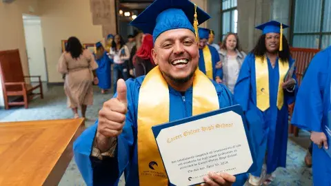 graduate wearing cap and gown holds up diploma with a thumbs up