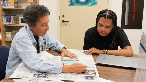 A person helping another person to read the newspaper. 