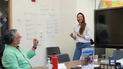 A teacher standing in front of a white board full of words. A student rising is their hand.