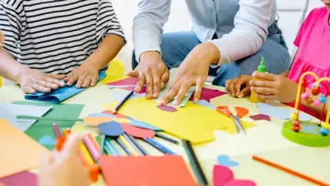 Hands are around a table creating art out of colorful materials. 