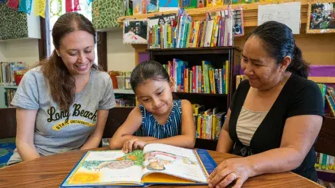 Two women sit at a table with a young girl in the middle. There is a picture book on the table and all three are smiling and reading the book. 