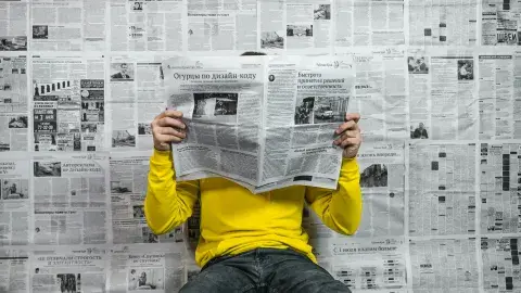 A man sitting on the floor reading a newspaper with newspaper wallpaper in the background