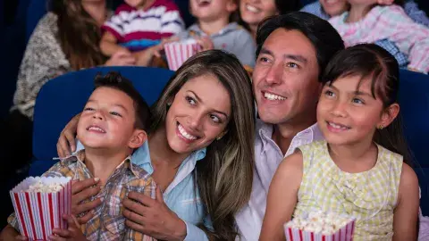 Two smiling parents and their children seated in a theater, enjoying a movie with popcorn in their laps