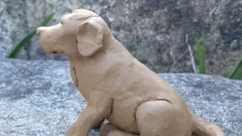 A clay dog facing sideways and sitting on its hind legs