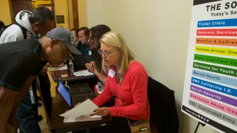 Seated woman in a red sweater at a table helping a standing man with paperwork.
