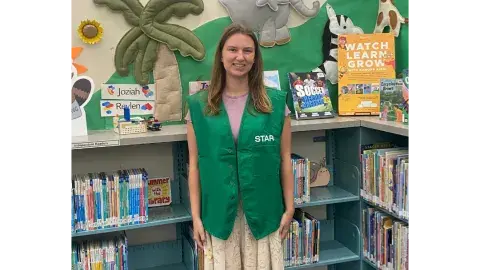 A young woman wearing a green "STAR" vest and a floral skirt standing in a children's library surrounded by books and colorful wall decorations.