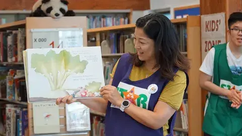 Smiling person wears a purple apron with the word STAR on it. The person is standing in front of library shelves and holding up a picture book.