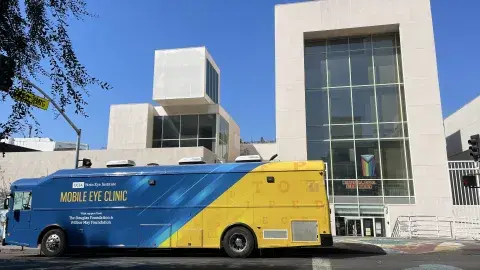 Blue and Yellow Mobile Eye Clinic Bus parked in fron tof a building. The blue sky is in the background.