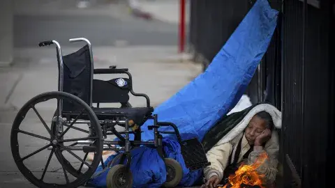 Person on the street huddled under tarp next to wheel chair with a fire burning