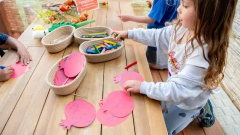 Three children sit at a wooden table with art supplies in small baskets, decorating pink paper fruits.