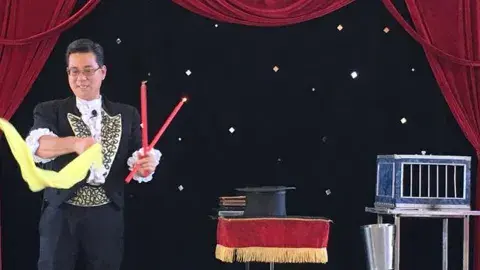 A magician holding wands and a scarf on stage next to a table with a top hat and a cage